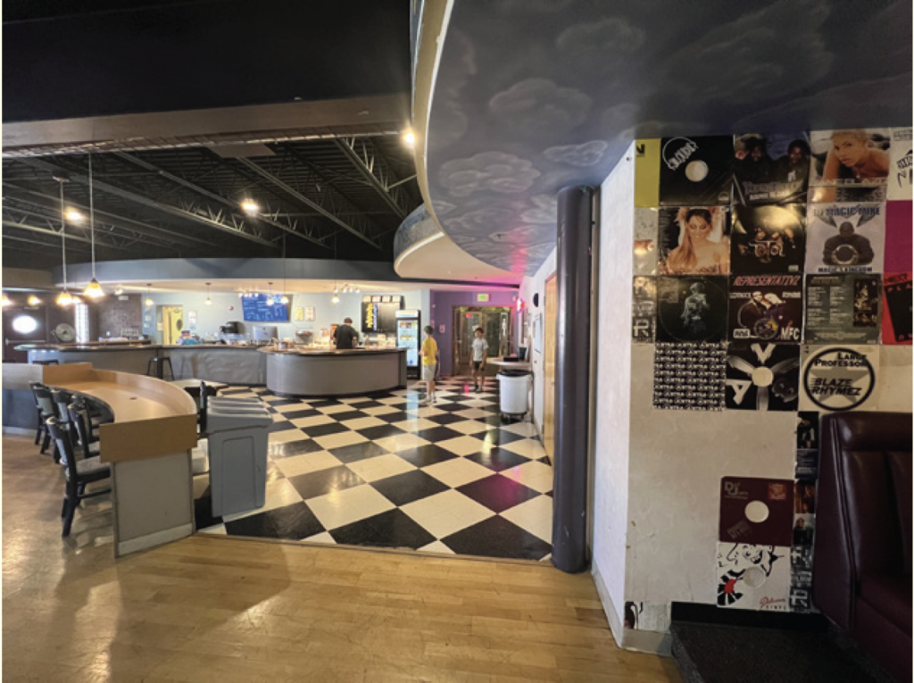 Interior of a diner with checkered flooring, booths, and bar seating on the left. A wall displaying various posters is on the right.